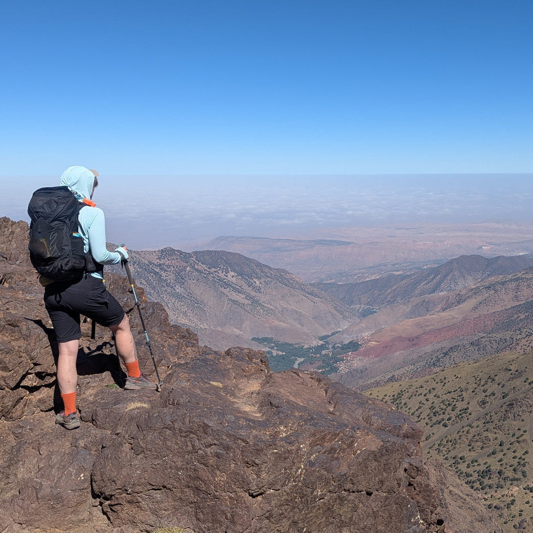 A hiker stands on a rocky mountain ridge overlooking a vast canyon valley under a clear blue sky, wearing a light blue top, black shorts, a black backpack, and orange socks.